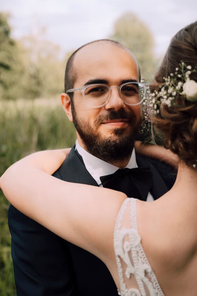 Groom smiling and looking into the camera while being hugged by his wife